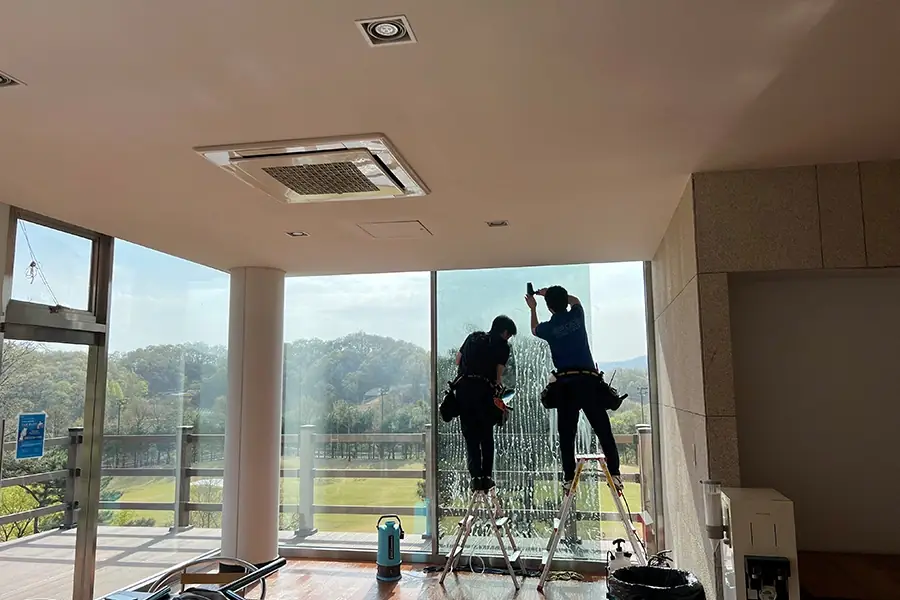 사다리 위에서 대형 창호에 필름을 부착하는 두 명의 시공기사